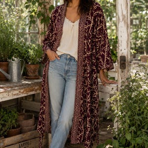 May include: A long, open-front burgundy kimono featuring a cream-colored branch pattern and a decorative geometric border along the edges. The garment is worn over a cream-colored camisole with lace trim, light-wash blue denim jeans, and brown sandals.