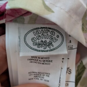 May include: A close-up of a fabric tag with the "laura ashley" logo in a white oval. The tag reads "MADE IN MEXICO" in English, French, and Spanish. The fabric has a floral pattern with pink and green accents.