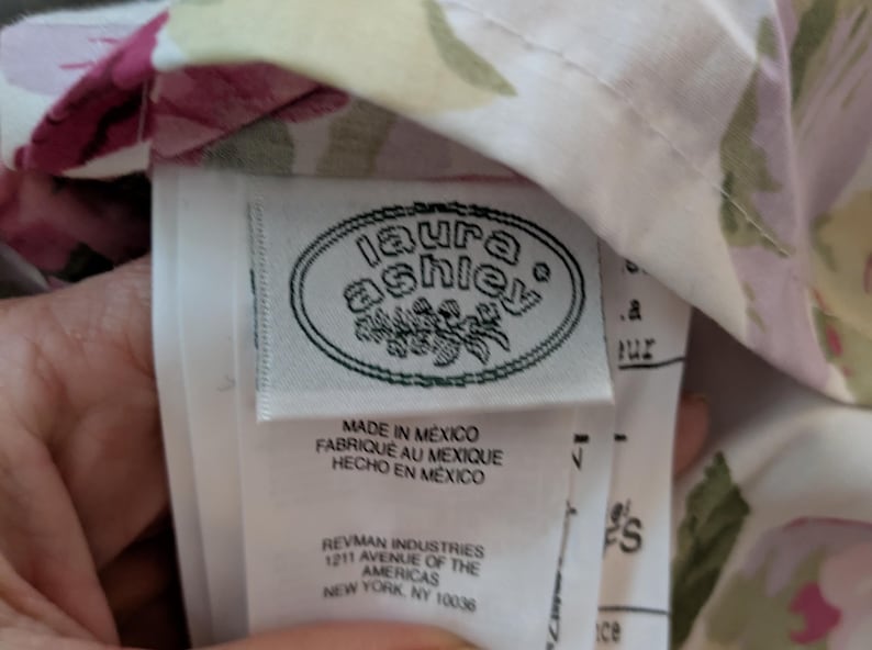 May include: A close-up of a fabric tag with the "laura ashley" logo in a white oval. The tag reads "MADE IN MEXICO" in English, French, and Spanish. The fabric has a floral pattern with pink and green accents.