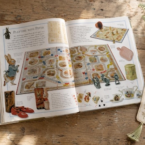 Könnte beinhalten: Ein aufgeschlagenes Buch mit Illustrationen und einem Brettspiel. Das Buch mit dem Titel "Playtime with Peter" enthält einen Lesezeichen mit Quaste. Weitere Artikel sind rote Schuhe und eine kleine Holzkiste.