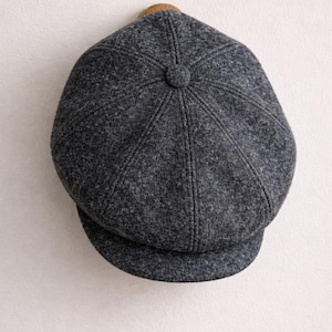 Puede incluir: Gorra gris de estilo newsboy con una corona redondeada y una visera pequeña y rígida. La gorra está hecha de una tela texturizada y tiene un botón en la parte superior. La gorra está colgada de un gancho de madera contra una pared blanca.
