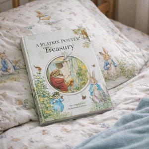 Peut inclure: Un livre intitulé « A Beatrix Potter Treasury » est posé sur un lit avec des taies d'oreiller assorties et une couverture bleu clair. La couverture du livre présente une illustration circulaire avec des personnages des histoires. La literie a un motif répété de lapins et d'éléments floraux.