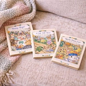 Könnte beinhalten: Drei Vintage-Kinderbücher mit Illustrationen von Hasen und anderen Tieren. Titel sind "Bunny Magic!", "A Tale Told Twice" und "Upside-Down Ears". Die Bücher liegen auf einer neutralen Oberfläche mit einer weichen, gemusterten Decke.