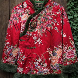 May include: A red jacket with a floral pattern and green fur trim. The jacket has a mandarin collar and frog closures. The design features flowers in pink, white, and yellow, set against a red background. The jacket is hanging on a wooden hanger.