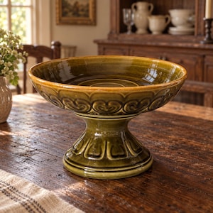 Puede incluir: Un cuenco con pedestal y esmalte verde oliva brillante. El cuenco presenta una cubeta ancha y poco profunda con una base robusta y acampanada. El exterior del cuenco y la base están decorados con un patrón en relieve de volutas y pétalos.