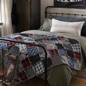 May include: A patchwork rag quilt featuring a variety of fabric squares in shades of red, blue, grey, and black, including plaid and patterned designs. The quilt has exposed frayed seams and is draped over the footboard of a black metal bed frame.