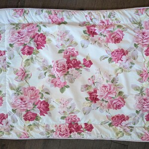 May include: A white floral quilt featuring a pattern of pink and red roses with green leaves. The quilt has a white border with a pink trim. The floral pattern is repeated across the entire surface of the quilt.
