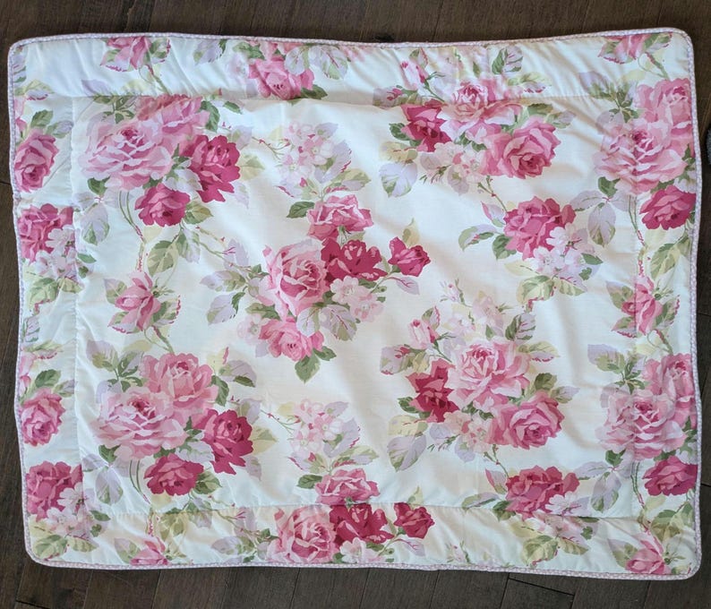 May include: A white floral quilt featuring a pattern of pink and red roses with green leaves. The quilt has a white border with a pink trim. The floral pattern is repeated across the entire surface of the quilt.
