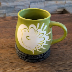 May include: A green ceramic mug with a white dove design. The mug sits on a stack of black coasters. The mug has a curved handle and a rounded body. The background is a wooden table and a stone fireplace.
