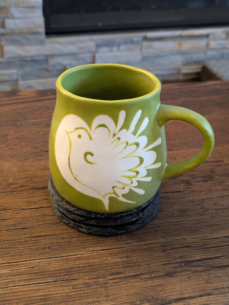 May include: A green ceramic mug with a white dove design. The mug sits on a stack of black coasters. The mug has a curved handle and a rounded body. The background is a wooden table and a stone fireplace.