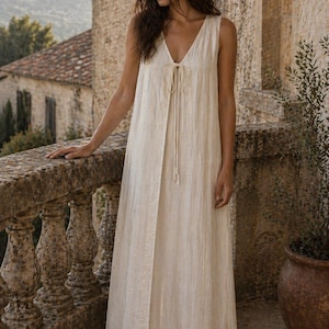 May include: A sleeveless, floor-length dress in a light beige color. The dress features a V-neckline with a tie detail and a front slit. The fabric has a subtle vertical texture. The dress is suitable for casual or formal wear.