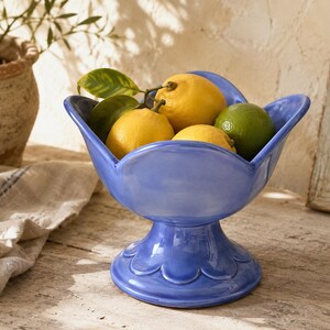May include: A glossy periwinkle blue ceramic pedestal bowl with a scalloped rim and base, filled with three bright yellow lemons with green leaves and one green lime. The bowl features a smooth, curved design with a wide, stable footed base.