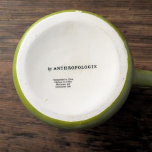 May include: A green ceramic mug with a white interior, viewed from the bottom. The text "by ANTHROPOLOGIE" is printed on the white surface. Additional text indicates the mug is handpainted in China and is microwave and dishwasher safe.