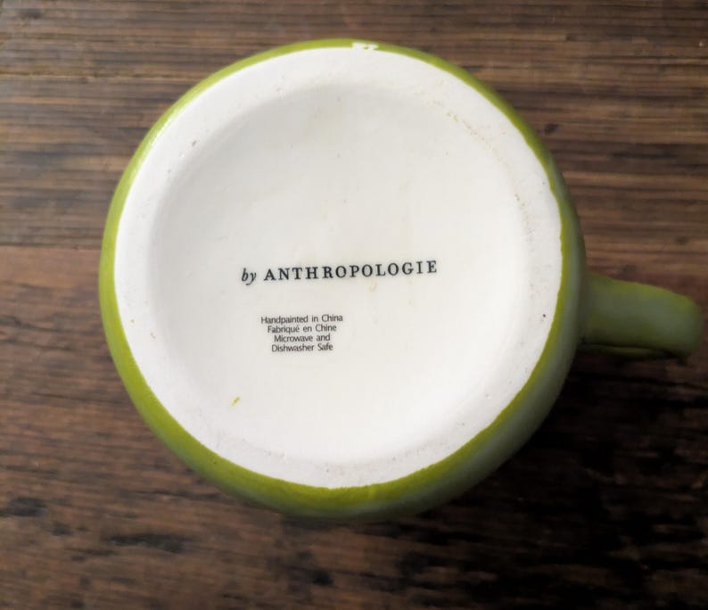 May include: A green ceramic mug with a white interior, viewed from the bottom. The text "by ANTHROPOLOGIE" is printed on the white surface. Additional text indicates the mug is handpainted in China and is microwave and dishwasher safe.