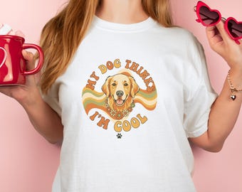 May include: White t-shirt with a retro graphic of a golden retriever wearing a flower collar, with the text "My Dog Thinks I'm Cool". The design features a paw print and a wavy, earthy color scheme.