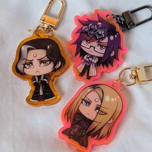 Op de afbeelding: Drie sleutelhangers met anime-personages. Elke sleutelhanger heeft een ander personage met gedetailleerde gezichtsuitdrukkingen en outfits. De sleutelhangers hebben goudkleurige sluitingen en zijn omlijnd met een feloranje rand.