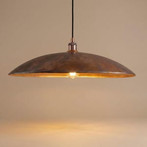 May include: A copper-colored pendant light fixture with a wide, shallow shade. The interior of the shade has a golden hue, and a single light bulb is visible. The fixture hangs from a black cord and has a copper-toned base.
