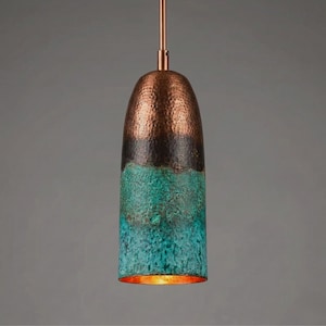 Op de afbeelding: Koperen hanglamp met een gehamerde textuur. De bovenkant is donkerbruin, overgaand in een turquoise en blauwgroene afwerking op de onderste helft. De binnenkant gloeit met warm licht, wat een unieke lamp creëert.