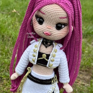 Puede incluir: Una mu&ntilde;eca de ganchillo con largo cabello magenta, grandes ojos negros y labios rosados. La mu&ntilde;eca viste una chaqueta blanca corta con botones dorados, un top negro y una falda blanca con ribete negro y detalles dorados. Texto AmiRo Dolls.