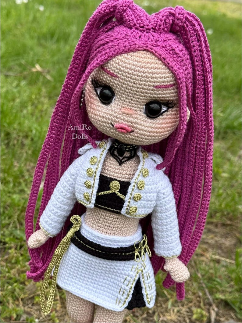 Puede incluir: Una mu&ntilde;eca de ganchillo con largo cabello magenta, grandes ojos negros y labios rosados. La mu&ntilde;eca viste una chaqueta blanca corta con botones dorados, un top negro y una falda blanca con ribete negro y detalles dorados. Texto AmiRo Dolls.