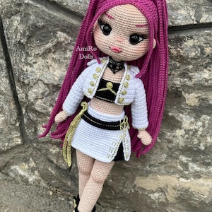 Puede incluir: Una mu&ntilde;eca de ganchillo hecha a mano con largo cabello magenta, que viste una chaqueta corta blanca con botones dorados, un top negro y blanco, una falda blanca a juego con detalles negros y zapatillas deportivas negras y blancas. El texto AmiRo Dolls es visible.