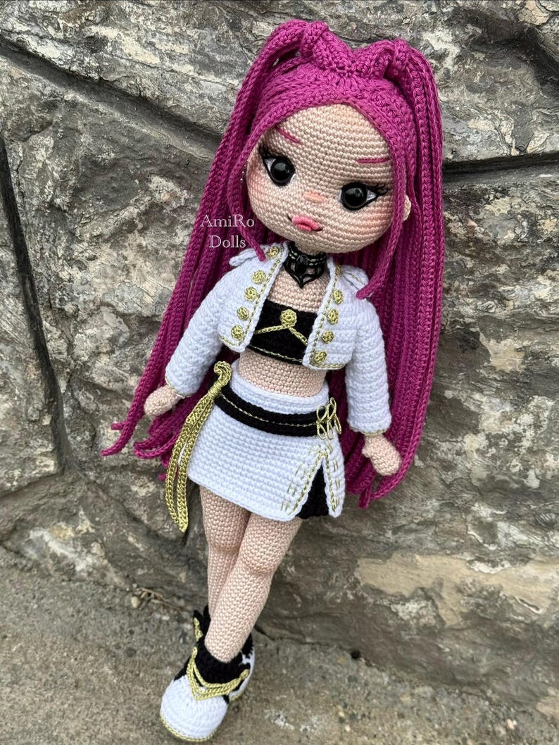 Puede incluir: Una mu&ntilde;eca de ganchillo hecha a mano con largo cabello magenta, que viste una chaqueta corta blanca con botones dorados, un top negro y blanco, una falda blanca a juego con detalles negros y zapatillas deportivas negras y blancas. El texto AmiRo Dolls es visible.