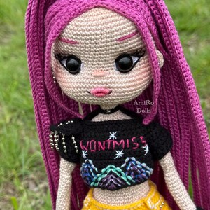 Puede incluir: Una mu&ntilde;eca de ganchillo con largo cabello magenta, vistiendo un top corto negro con el texto WONTMISS y un gr&aacute;fico de monta&ntilde;a, pantalones amarillos con una cadena plateada y guantes negros sin dedos. La mu&ntilde;eca tiene grandes ojos negros.