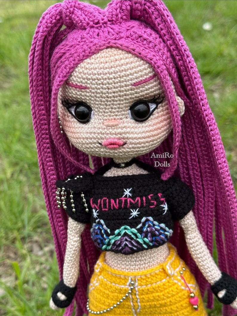 Puede incluir: Una mu&ntilde;eca de ganchillo con largo cabello magenta, vistiendo un top corto negro con el texto WONTMISS y un gr&aacute;fico de monta&ntilde;a, pantalones amarillos con una cadena plateada y guantes negros sin dedos. La mu&ntilde;eca tiene grandes ojos negros.