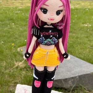 Puede incluir: Una mu&ntilde;eca de ganchillo con largo cabello magenta, que viste un top corto negro con el texto WONT MISS y un gr&aacute;fico de monta&ntilde;a, una falda amarilla con una cadena dorada, guantes negros sin dedos y botas negras con detalles en rosa y blanco.