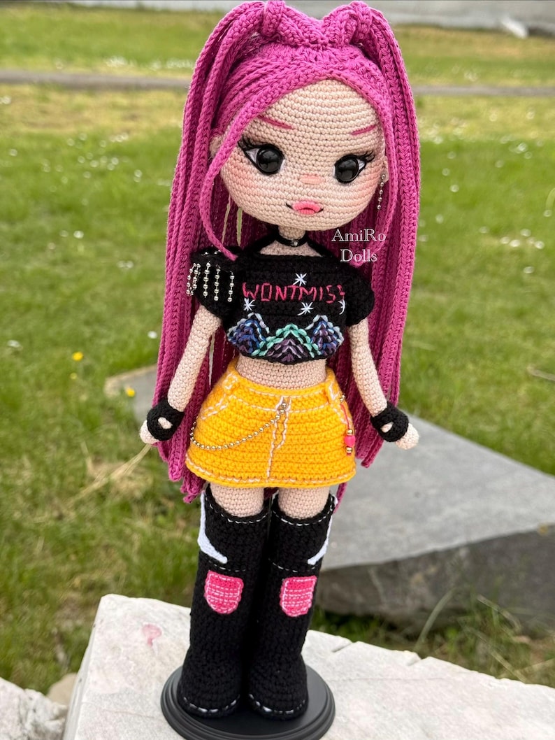 Puede incluir: Una mu&ntilde;eca de ganchillo con largo cabello magenta, que viste un top corto negro con el texto WONT MISS y un gr&aacute;fico de monta&ntilde;a, una falda amarilla con una cadena dorada, guantes negros sin dedos y botas negras con detalles en rosa y blanco.