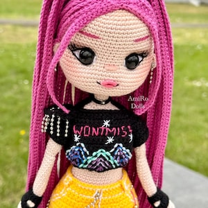 Puede incluir: Una mu&ntilde;eca de ganchillo con largo cabello rosa brillante, que viste un top corto negro con el texto "WONTMISS" y un gr&aacute;fico de monta&ntilde;a, combinado con una falda amarilla. La mu&ntilde;eca tiene grandes ojos negros, labios rosas, guantes negros sin dedos y una cadena plateada.