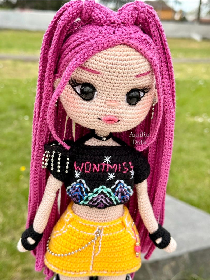 Puede incluir: Una mu&ntilde;eca de ganchillo con largo cabello rosa brillante, que viste un top corto negro con el texto "WONTMISS" y un gr&aacute;fico de monta&ntilde;a, combinado con una falda amarilla. La mu&ntilde;eca tiene grandes ojos negros, labios rosas, guantes negros sin dedos y una cadena plateada.