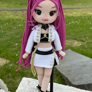 Puede incluir: Una mu&ntilde;eca de ganchillo con largo cabello magenta, que viste una chaqueta corta blanca con botones dorados, un top negro y una falda blanca con cintur&oacute;n negro. La mu&ntilde;eca tiene grandes ojos negros, labios rosas y zapatos blancos y negros. Texto: AmiRo Dolls.