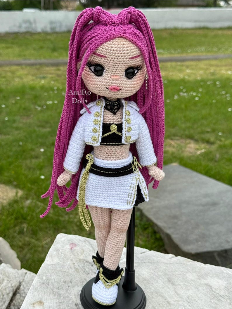 Puede incluir: Una mu&ntilde;eca de ganchillo con largo cabello magenta, que viste una chaqueta corta blanca con botones dorados, un top negro y una falda blanca con cintur&oacute;n negro. La mu&ntilde;eca tiene grandes ojos negros, labios rosas y zapatos blancos y negros. Texto: AmiRo Dolls.