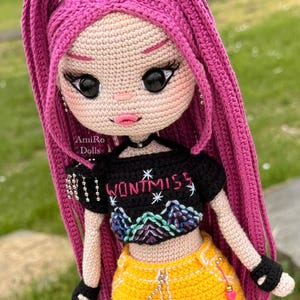 Puede incluir: Una mu&ntilde;eca de ganchillo con largo cabello rosa vibrante, que viste un top corto negro con el texto "WONT MISS" y un gr&aacute;fico de monta&ntilde;a, combinado con una minifalda amarilla brillante con detalle de cadena plateada y guantes negros.