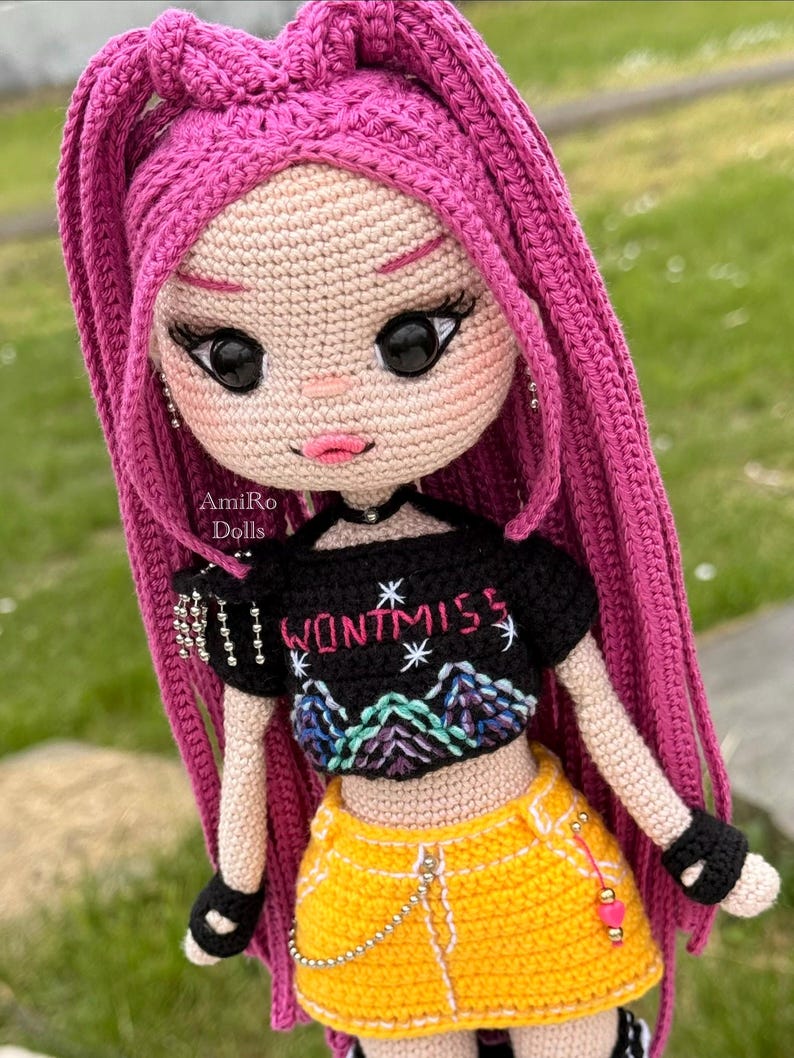 Puede incluir: Una mu&ntilde;eca de ganchillo con largo cabello rosa vibrante, que viste un top corto negro con el texto "WONT MISS" y un gr&aacute;fico de monta&ntilde;a, combinado con una minifalda amarilla brillante con detalle de cadena plateada y guantes negros.