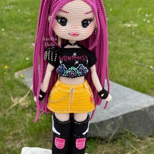 Puede incluir: Una mu&ntilde;eca tejida a mano con largo cabello rosa vibrante, que viste un top corto negro con el texto WONT MISS y un gr&aacute;fico de monta&ntilde;a, una minifalda amarilla con una cadena plateada y botas negras hasta la rodilla con detalles.