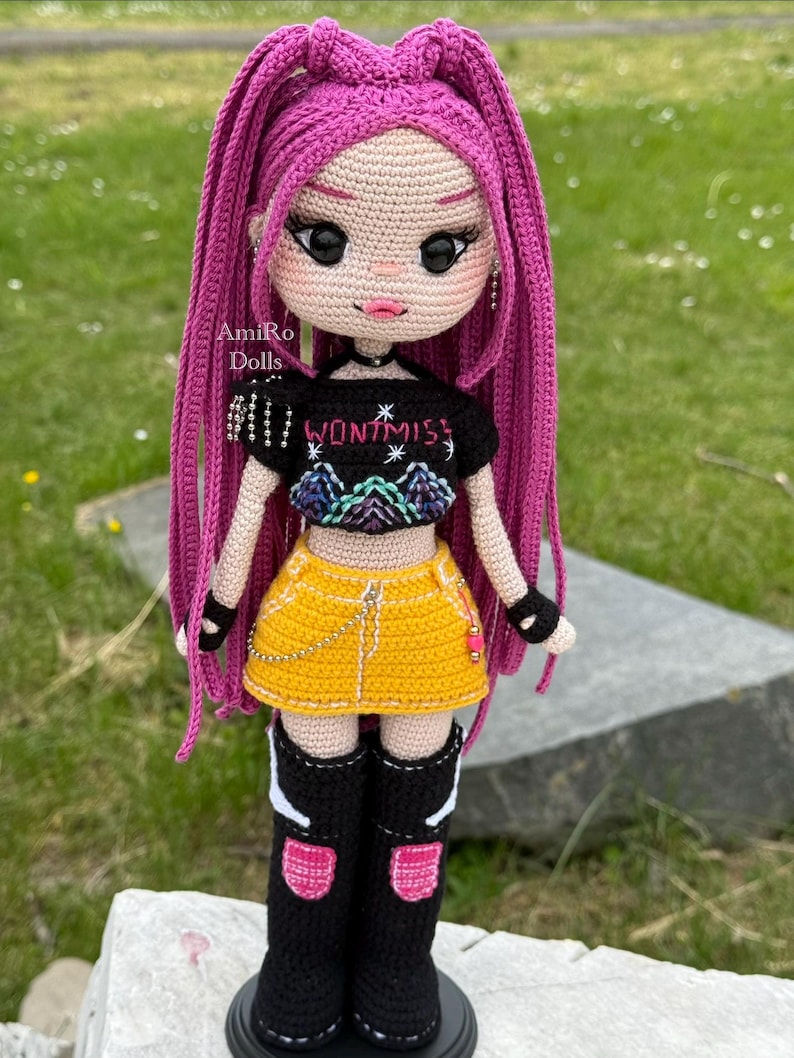 Puede incluir: Una mu&ntilde;eca tejida a mano con largo cabello rosa vibrante, que viste un top corto negro con el texto WONT MISS y un gr&aacute;fico de monta&ntilde;a, una minifalda amarilla con una cadena plateada y botas negras hasta la rodilla con detalles.