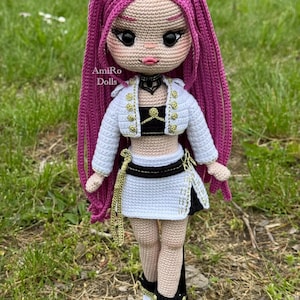 Puede incluir: Una mu&ntilde;eca de ganchillo con largo cabello magenta, que viste una chaqueta corta blanca con botones dorados, un top negro, una falda blanca con cinturilla negra y zapatillas blancas con detalles dorados. El texto AmiRo Dolls es visible en el pecho.