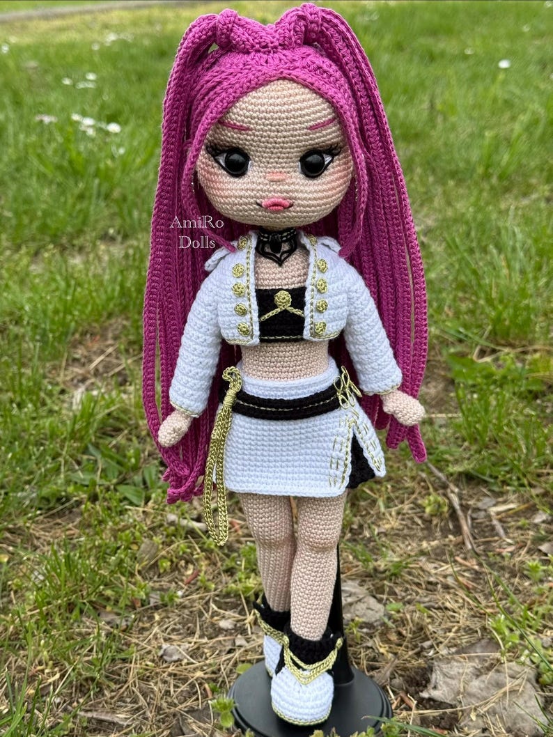 Puede incluir: Una mu&ntilde;eca de ganchillo con largo cabello magenta, que viste una chaqueta corta blanca con botones dorados, un top negro, una falda blanca con cinturilla negra y zapatillas blancas con detalles dorados. El texto AmiRo Dolls es visible en el pecho.