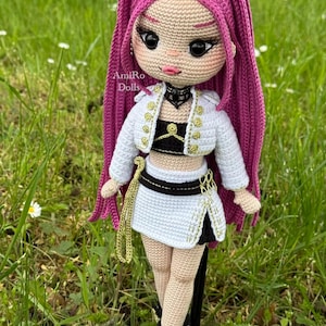 Puede incluir: Una mu&ntilde;eca de ganchillo hecha a mano con largo cabello magenta, que viste una chaqueta corta blanca con botones dorados, un top negro y una falda blanca con detalles negros. La mu&ntilde;eca tiene grandes ojos oscuros, una gargantilla negra y zapatillas blancas.