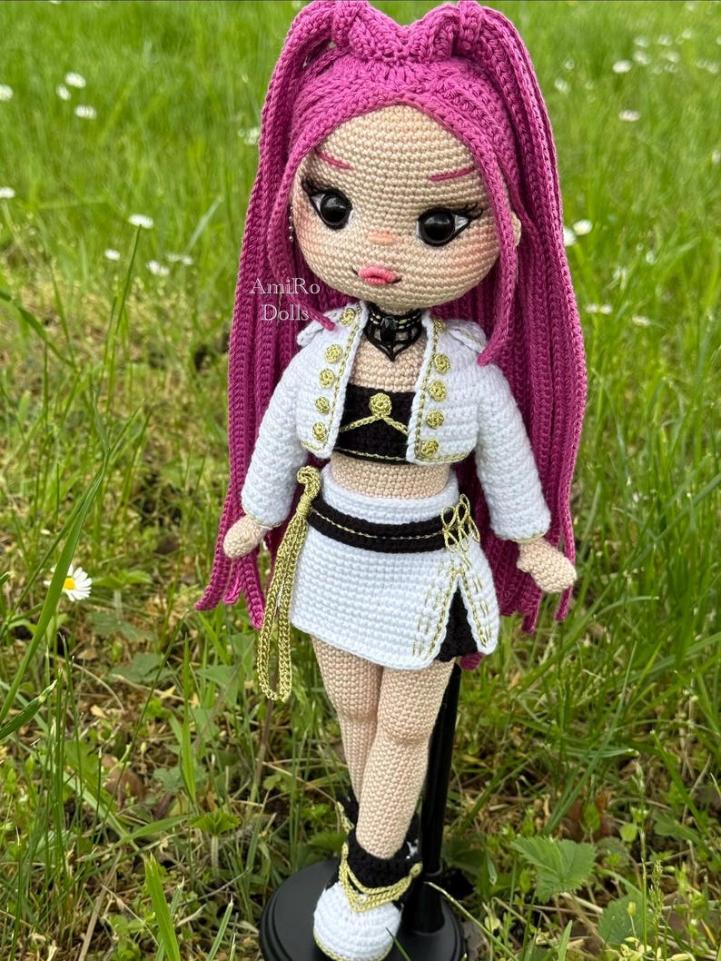 Puede incluir: Una mu&ntilde;eca de ganchillo hecha a mano con largo cabello magenta, que viste una chaqueta corta blanca con botones dorados, un top negro y una falda blanca con detalles negros. La mu&ntilde;eca tiene grandes ojos oscuros, una gargantilla negra y zapatillas blancas.
