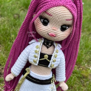 Puede incluir: Una mu&ntilde;eca tejida a mano con largo cabello magenta, que viste una chaqueta corta blanca con botones dorados, un top negro y una falda blanca con detalles negros. La mu&ntilde;eca tiene ojos negros grandes, labios rosados y una gargantilla negra. Texto: AmiRo Dolls.