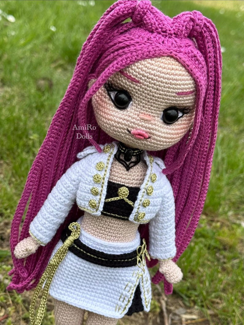 Puede incluir: Una mu&ntilde;eca tejida a mano con largo cabello magenta, que viste una chaqueta corta blanca con botones dorados, un top negro y una falda blanca con detalles negros. La mu&ntilde;eca tiene ojos negros grandes, labios rosados y una gargantilla negra. Texto: AmiRo Dolls.
