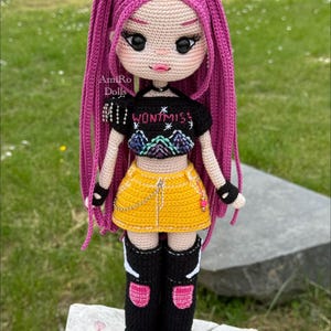 Puede incluir: Una mu&ntilde;eca de ganchillo con largo cabello magenta, que viste un top corto negro con el texto "WONTMISS" y un gr&aacute;fico de monta&ntilde;a, una minifalda amarilla con una cadena plateada, guantes negros sin dedos y botas altas negras con detalles rosas.