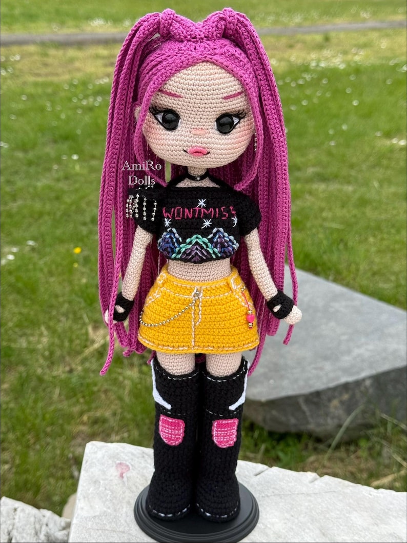 Puede incluir: Una mu&ntilde;eca de ganchillo con largo cabello magenta, que viste un top corto negro con el texto "WONTMISS" y un gr&aacute;fico de monta&ntilde;a, una minifalda amarilla con una cadena plateada, guantes negros sin dedos y botas altas negras con detalles rosas.