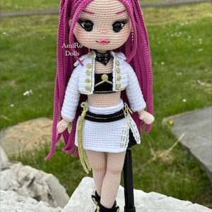 Puede incluir: Una mu&ntilde;eca de ganchillo hecha a mano con largo cabello magenta, que viste una chaqueta corta blanca con botones dorados, un top negro y una falda blanca con detalles dorados. La mu&ntilde;eca tiene grandes ojos negros, labios rosas y zapatillas. Texto: AmiRo Dolls.