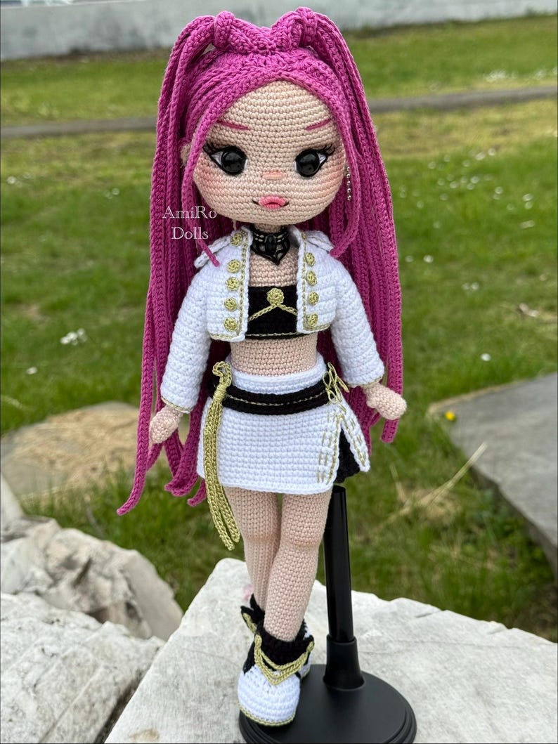 Puede incluir: Una mu&ntilde;eca de ganchillo hecha a mano con largo cabello magenta, que viste una chaqueta corta blanca con botones dorados, un top negro y una falda blanca con detalles dorados. La mu&ntilde;eca tiene grandes ojos negros, labios rosas y zapatillas. Texto: AmiRo Dolls.