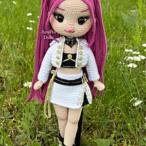 Puede incluir: Una mu&ntilde;eca de ganchillo con largo cabello magenta, que viste una chaqueta corta blanca con botones dorados, un top negro y una falda blanca con detalles negros. La mu&ntilde;eca tiene grandes ojos negros, labios rosas y una gargantilla negra. El texto AmiRo Dolls es visible.