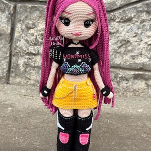 Puede incluir: Una mu&ntilde;eca de ganchillo hecha a mano con largo cabello rosa vibrante, que viste un top corto negro con el texto WONT MISS y un gr&aacute;fico de monta&ntilde;a, una minifalda amarilla brillante con una cadena plateada y botas negras altas con detalles.